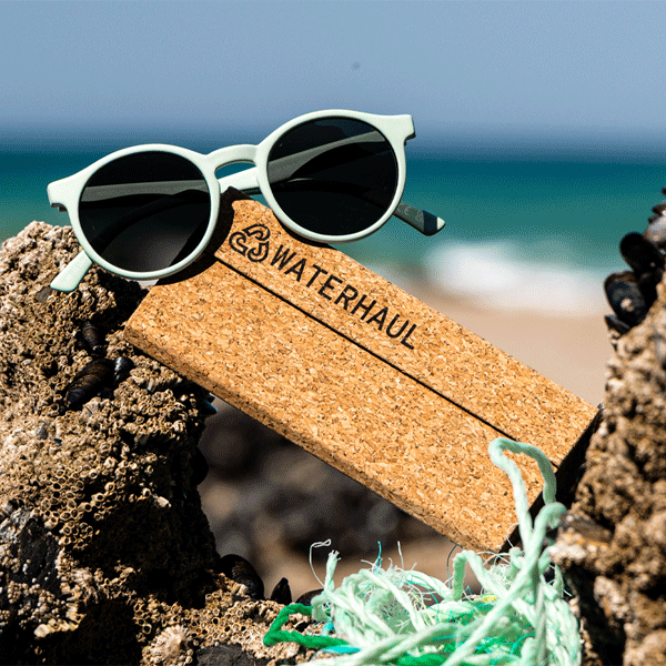 Sunglasses on a cork glasses case with 'WATERHAUL' branding, set against a beach background.