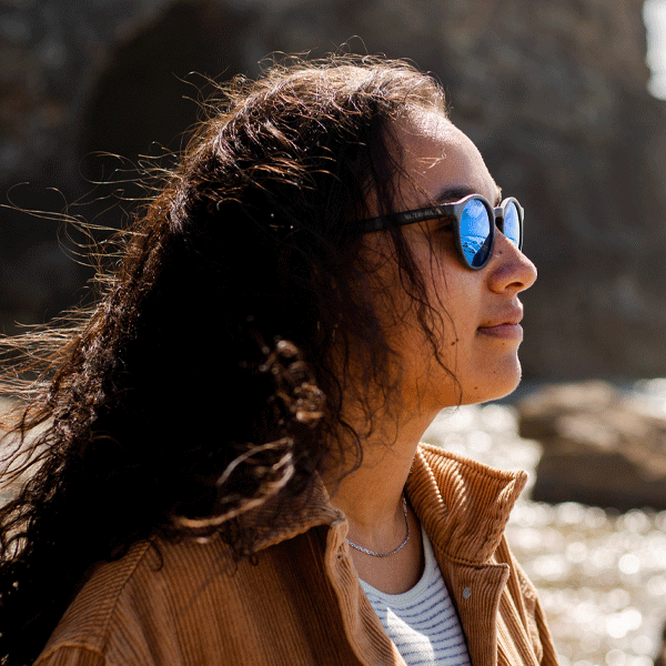 Woman wearing sunglasses with blue reflective lenses, standing outdoors near water.