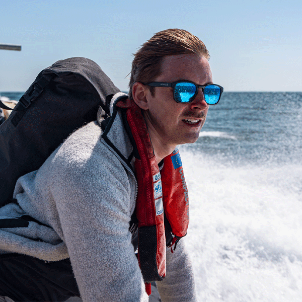 Man on a boat wearing sunglasses and a life jacket with a clear blue sky and ocean background.