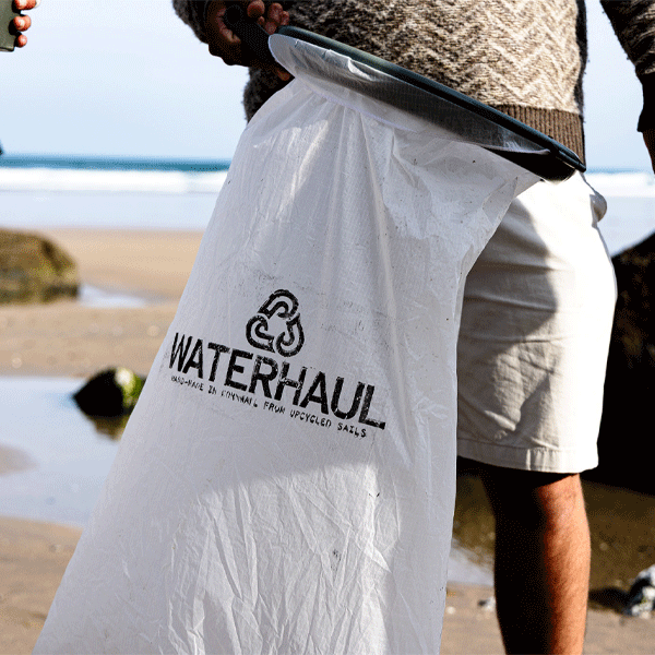 Person holding a white trash bag with 'Waterhaul' branding on a beach.